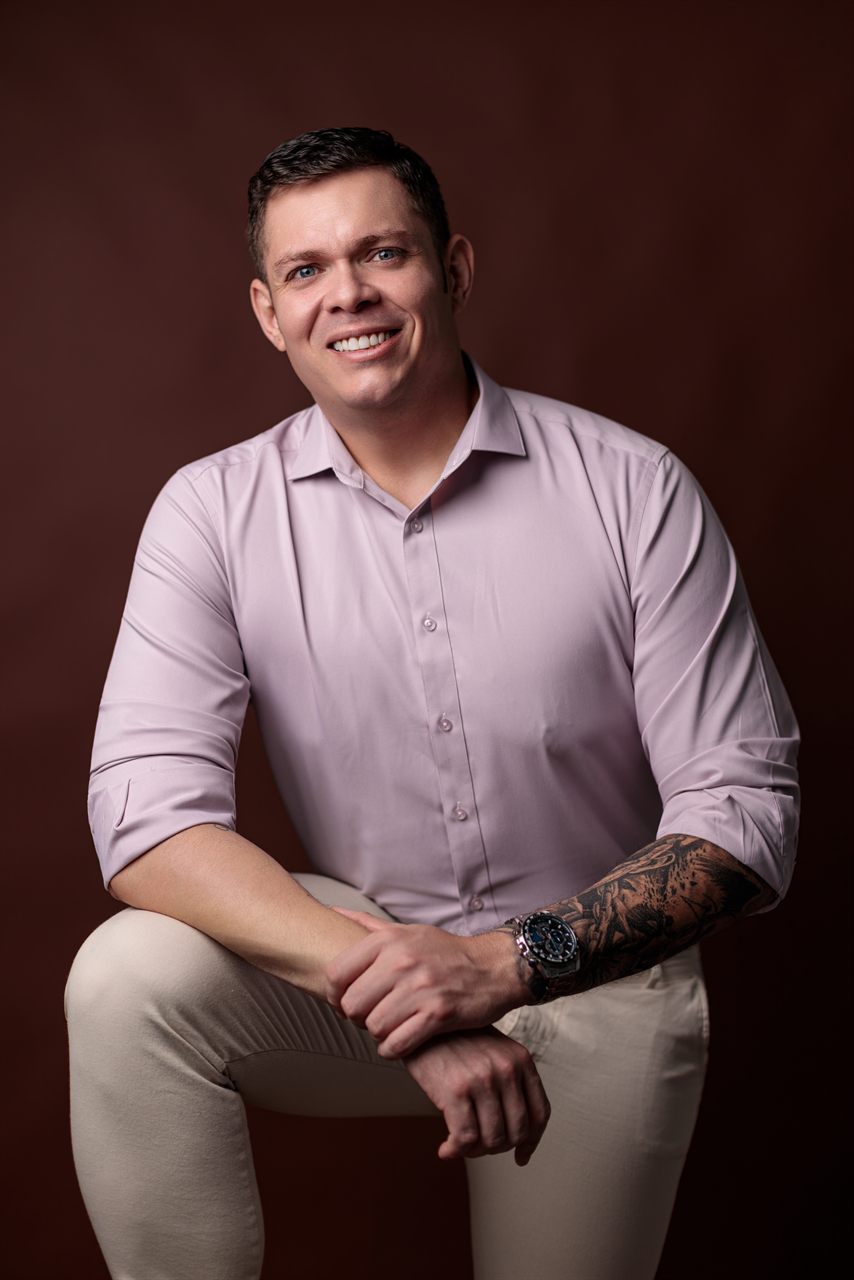 Renan Souza sorrindo, de camisa rosa-clara, em fundo escuro